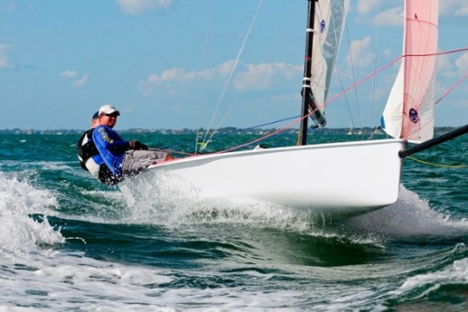Vx One sailing boat cutting across open water under a steady breeze. The lightweight hull sits low on the waves while the tall mast and clean sail plan show its racing focus. Crew members balance on the edge, trimming sails for speed, with sunlight reflecting off the water around the boat.